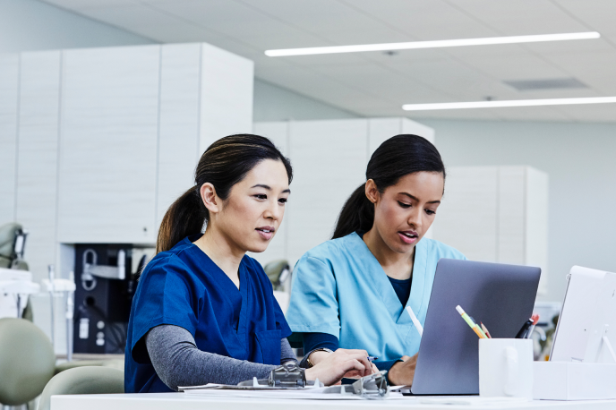 Duas enfermeiras sentadas em frente aos seus computadores, fazendo anotações