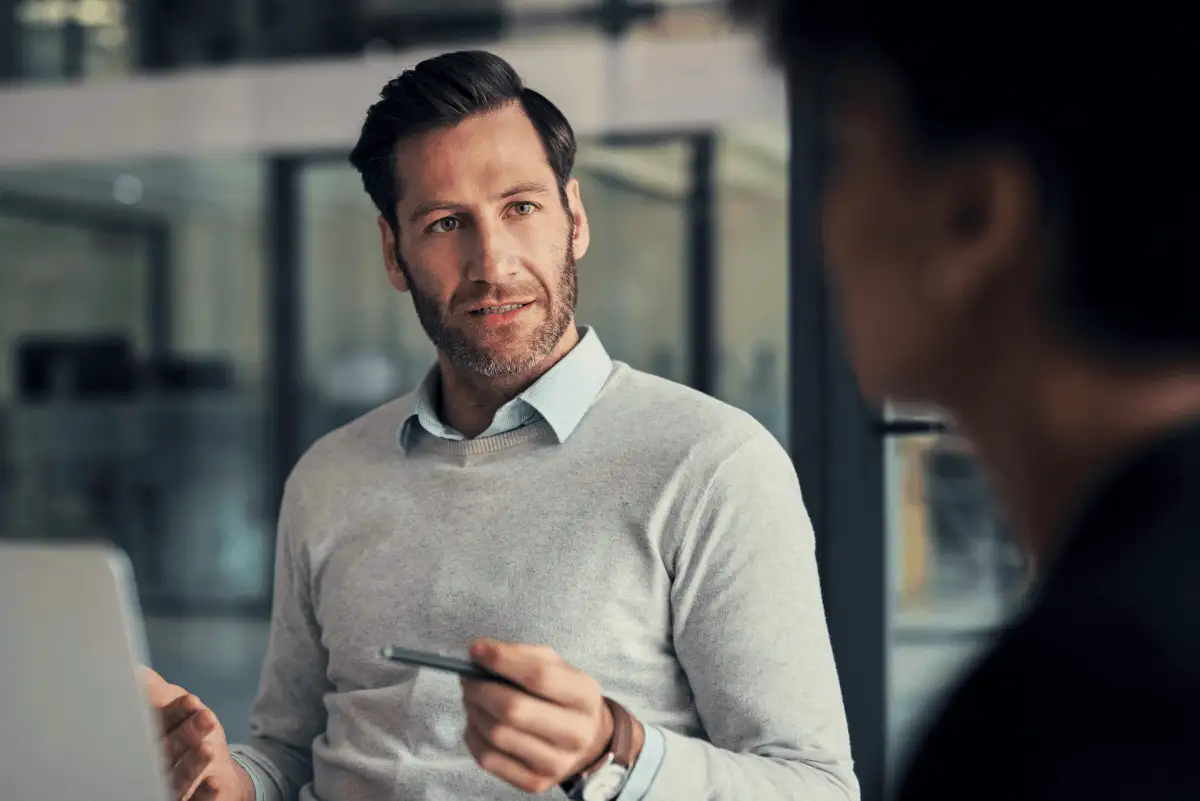 A man in an office holding a pen and talking to a coworker